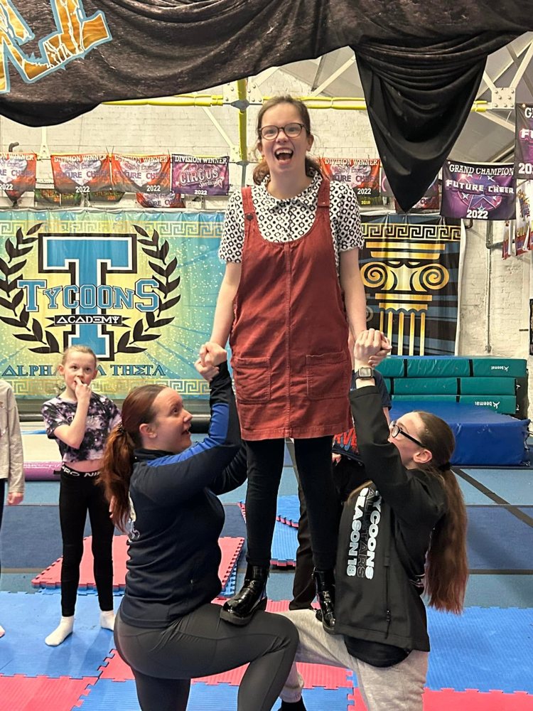 A smiling Tycoons SEND athlete being lifted in a stunt by two coaches